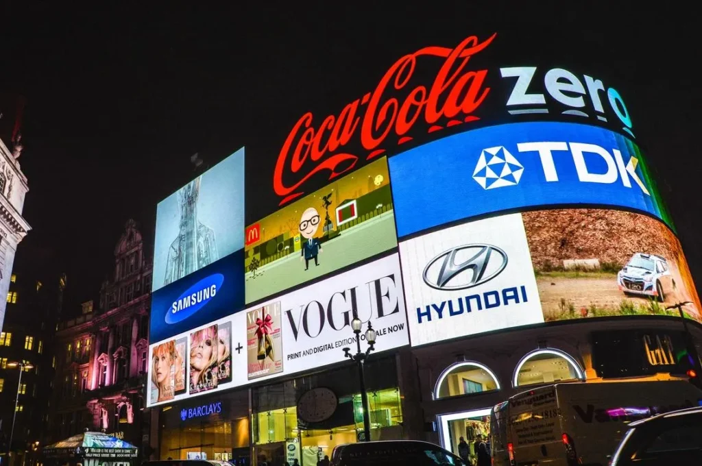 Illuminated digital billboards in a city at night displaying major global brands including Coca-Cola, Samsung, McDonald’s, Vogue, Hyundai and TDK