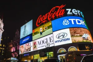 Illuminated digital billboards in a city at night displaying major global brands including Coca-Cola, Samsung, McDonald’s, Vogue, Hyundai and TDK