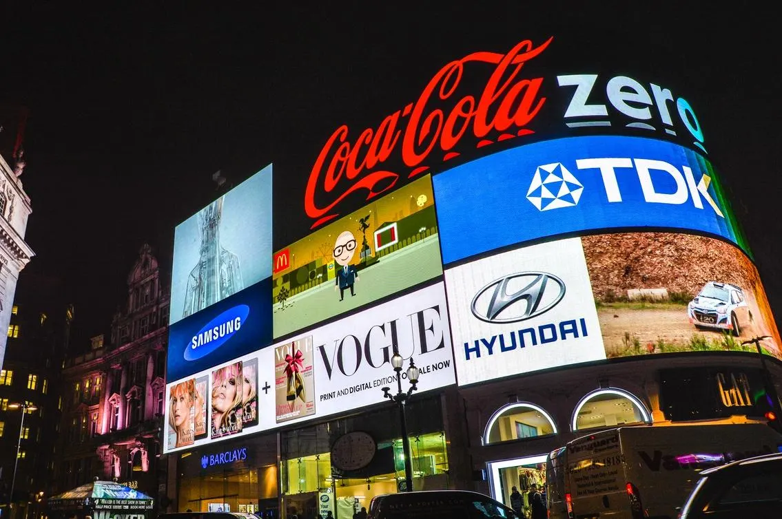 Illuminated digital billboards in a city at night displaying major global brands including Coca-Cola, Samsung, McDonald’s, Vogue, Hyundai and TDK