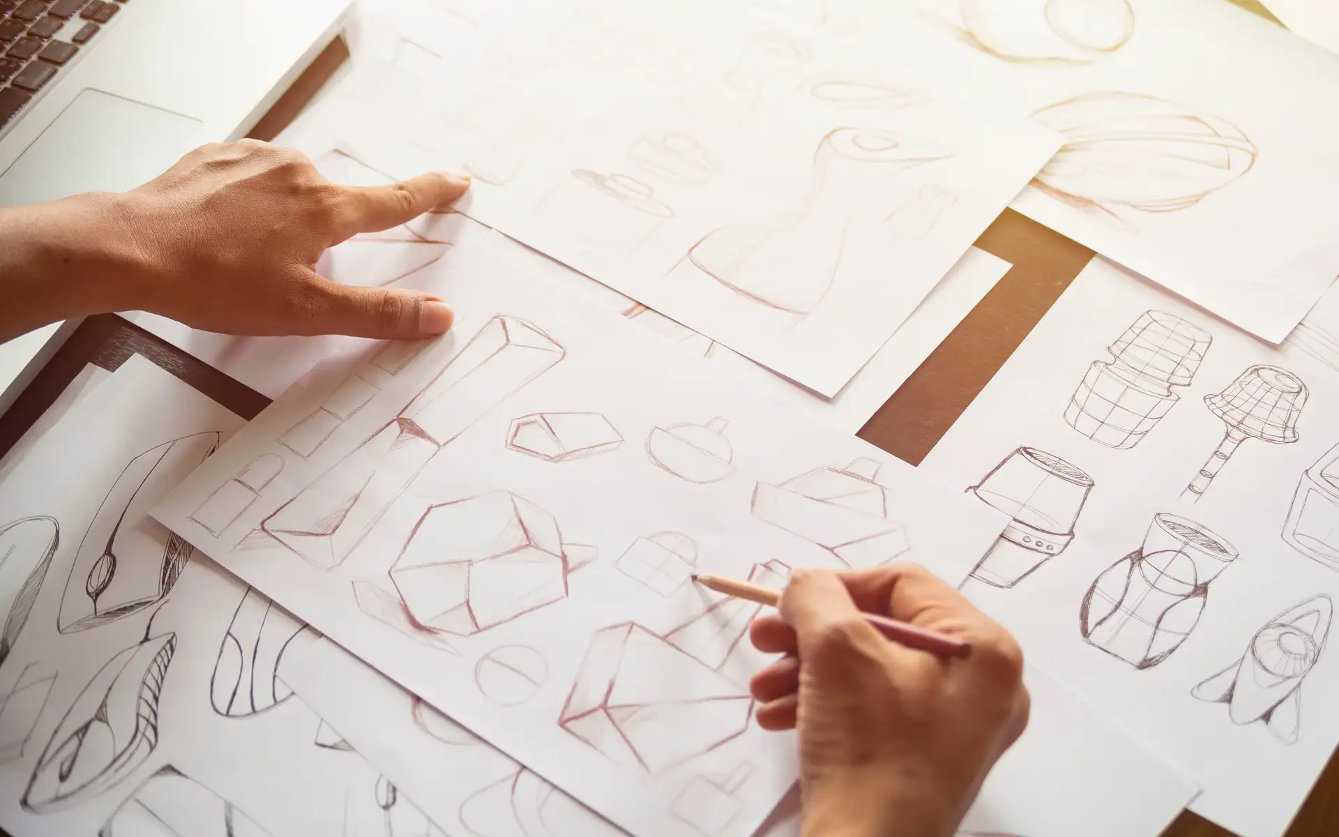 Hands sketching product concepts and geometric designs on paper alongside laptop.